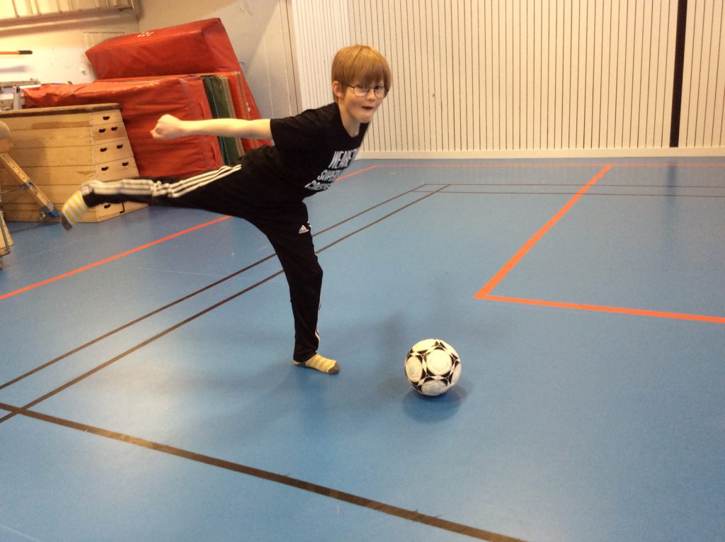 Noel står i en idrottshall med ett höjt ben och ska precis sparka på en fotboll.