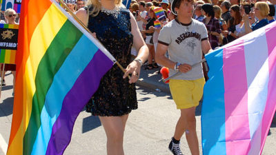 Två personer går med varsin flagga. Den ena flaggan har regnbågens färger. Den andra är transflaggan som är blå rosa och vit.