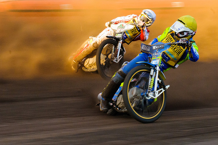 Två speedwaymotorcyklar åker på en grusbana. Båda förarna lutar cyklarna väldigt mycket och det sprutar sand. Båda har mycket skydd och stora hjälmar på sig.