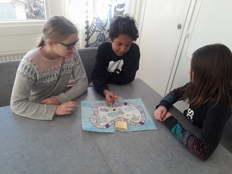 Tre elever sitter runt ett bord i ett klassrum och spelar ett spel. Spelet föreställer en stig runt några träd.