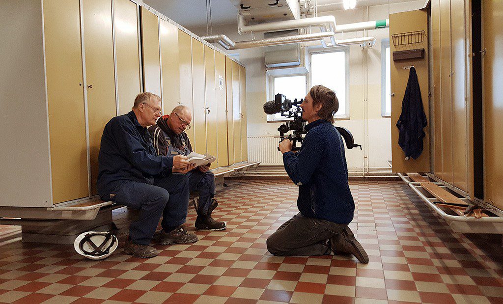 Ett omklädningsrum. På en bänk framför en rad med skåp sitter två äldre män. De har blå overaller på sig och tittar på ett foto. Framför dem sitter Nils Petter Löfstedt på knä och filmar dem med sin kamera.