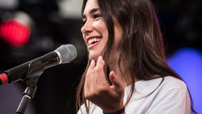 Dua Lipa står på en scen framför en mikrofon. Hon har långt brunt hår och en vit t-shirt. Hon ler stort.