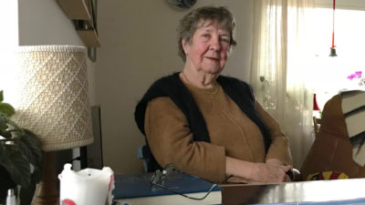 Dorothy Samuelsson har en svart väst över en brun tröja. Hon sitter vid ett bord i sitt vardagsrum.