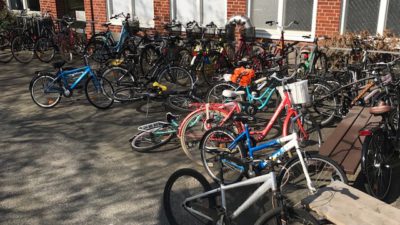 En cykelparkering intill en vägg som är helt full av cyklar som ligger välta på marken och många som står utanför cykelställen.