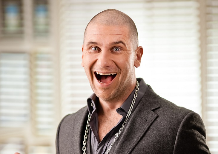 Markoolio ler med en jättestor öppen mun. Han har kavaj och skjorta på sig, och en stor kedja runt halsen. Han har snaggat hår och glada ögon. Han ser lite galen ut.