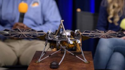 På ett bord står en liten modell av en maskin. Den har tre ben och en metallkropp. På sidorna går det ut två saker som ser lite ut som vingar.