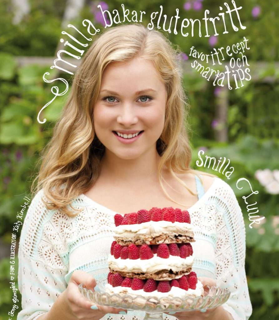 En bild på boken. Smilla står utomhus på sommaren. Hon ler och håller i en tårta som består av flera lager maräng med hela hallon och grädde emellan.