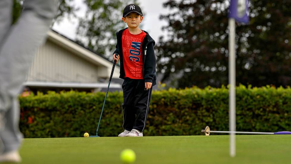 Erik står på golfbanan. Han har keps och träningsbyxor på sig. Han har klubben i handen. Han tittar bort mot bollen som han har puttat.