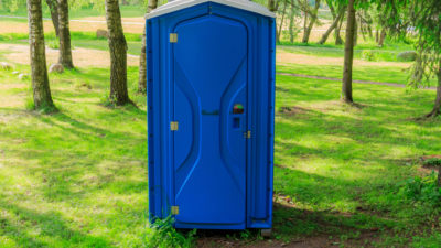 En bajamaja står på en gräsmatta. Bakom syns flera träd. Bajamajan ser ut som en hytt av plast.